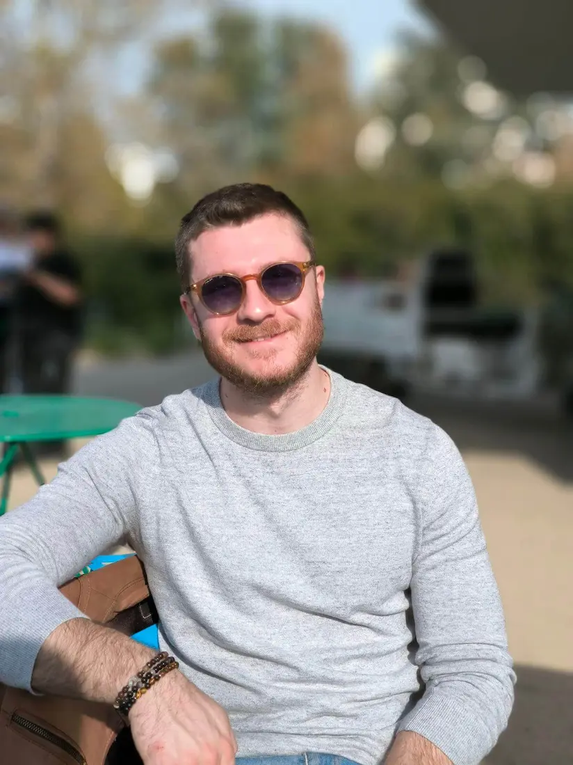 Alex Moudatsos wearing sunglasses sits at an outdoor table, enjoying the ambiance of a sunny day.